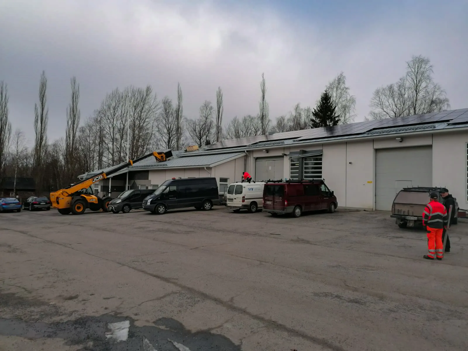 Työntekijät asentavat aurinkovoimalan paneeleja rakennuksen katolle nosturin ja pakettiautojen avustuksella.